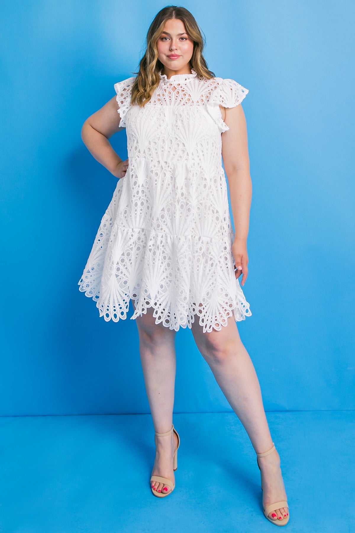 A woman is standing confidently, wearing a white, intricately patterned dress with ruffled sleeves. She is posed against a solid blue background, complemented by beige high-heeled sandals.