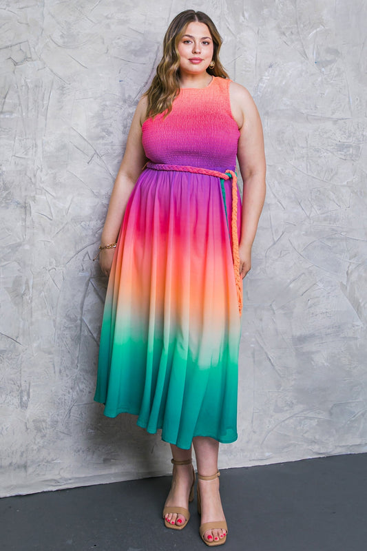 A woman stands confidently in a vibrant, multicolored dress transitioning from pink to orange to green. She poses against a textured gray backdrop, showcasing her stylish, open-toe heels.