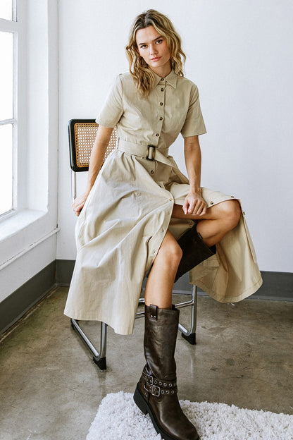 A person sits on a chair, wearing a tan, button-up dress with a belt. They're styled with tall brown boots, against a minimalistic, light-filled background featuring concrete and white walls.