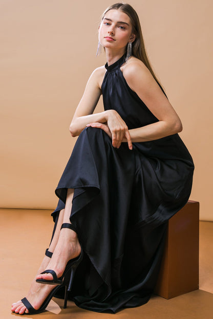 A woman sits elegantly on a brown block, wearing a black halter dress and high-heeled sandals. Her expression is serene, with long hair cascading over her shoulders against a soft beige background.