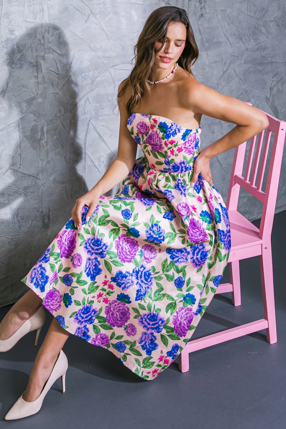 A woman in a floral, strapless dress sits on a pink chair, with one hand on her hip and the other resting beside her. The background features a textured, gray wall.