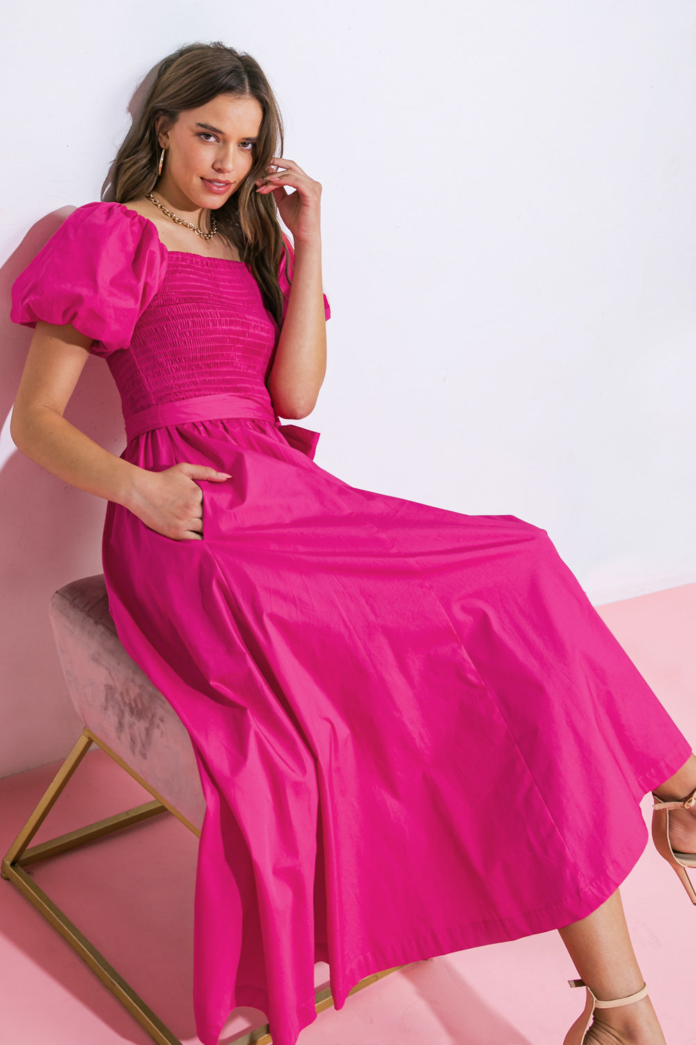 A woman wears a bright pink dress with puffed sleeves, sitting on a gold-accented stool. She rests one hand casually on her leg, against a white wall and pink floor.