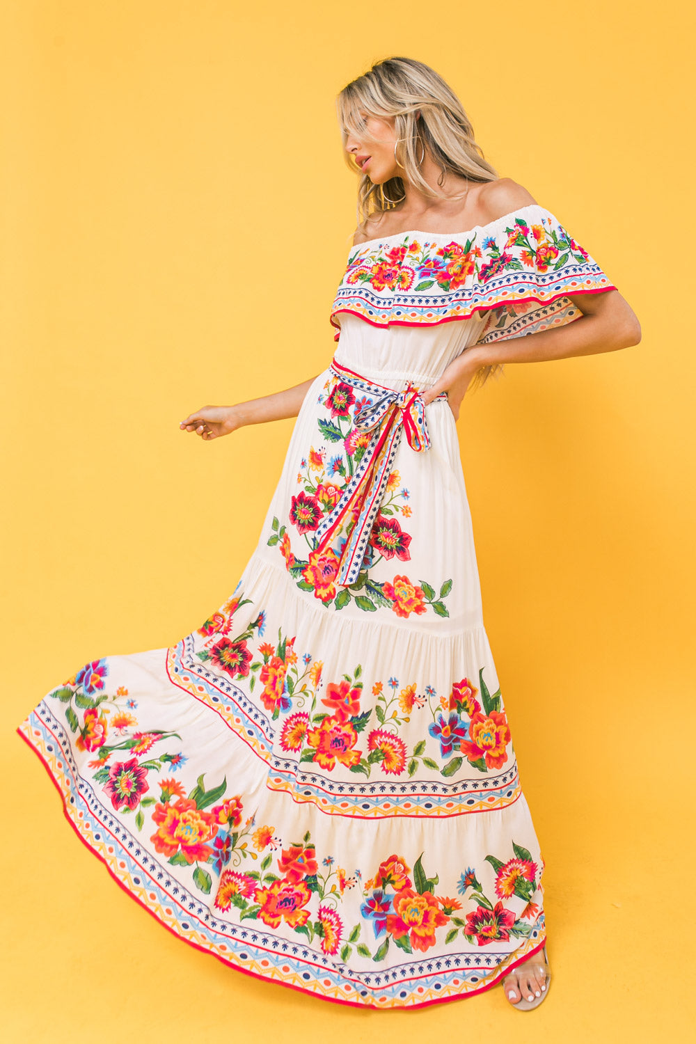 A woman in a vibrant, floral-patterned off-the-shoulder maxi dress spins joyfully against a bright yellow background, showcasing the dress's colorful embroidery and flowing fabric.