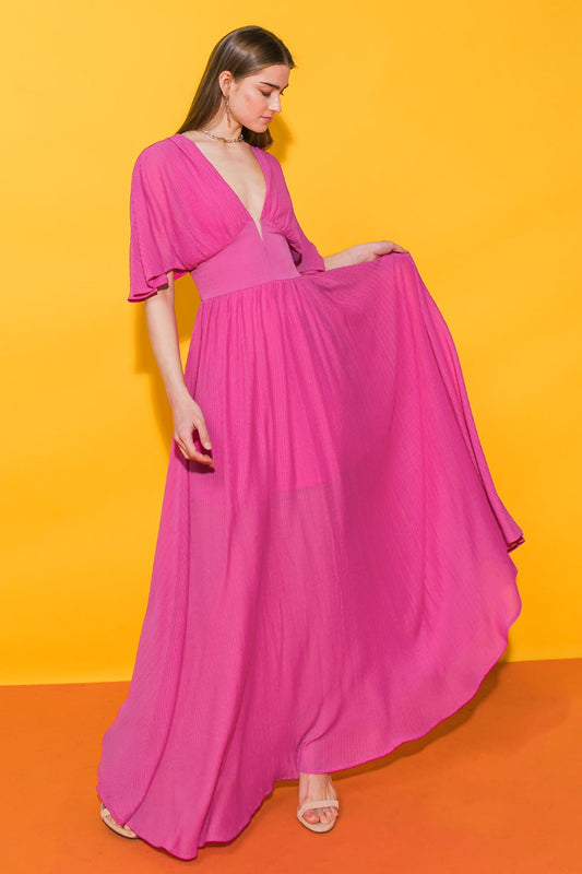 A woman in a flowing pink dress gracefully holds the fabric while standing against a bright yellow wall, complemented by a warm orange floor, creating a vibrant, cheerful atmosphere.