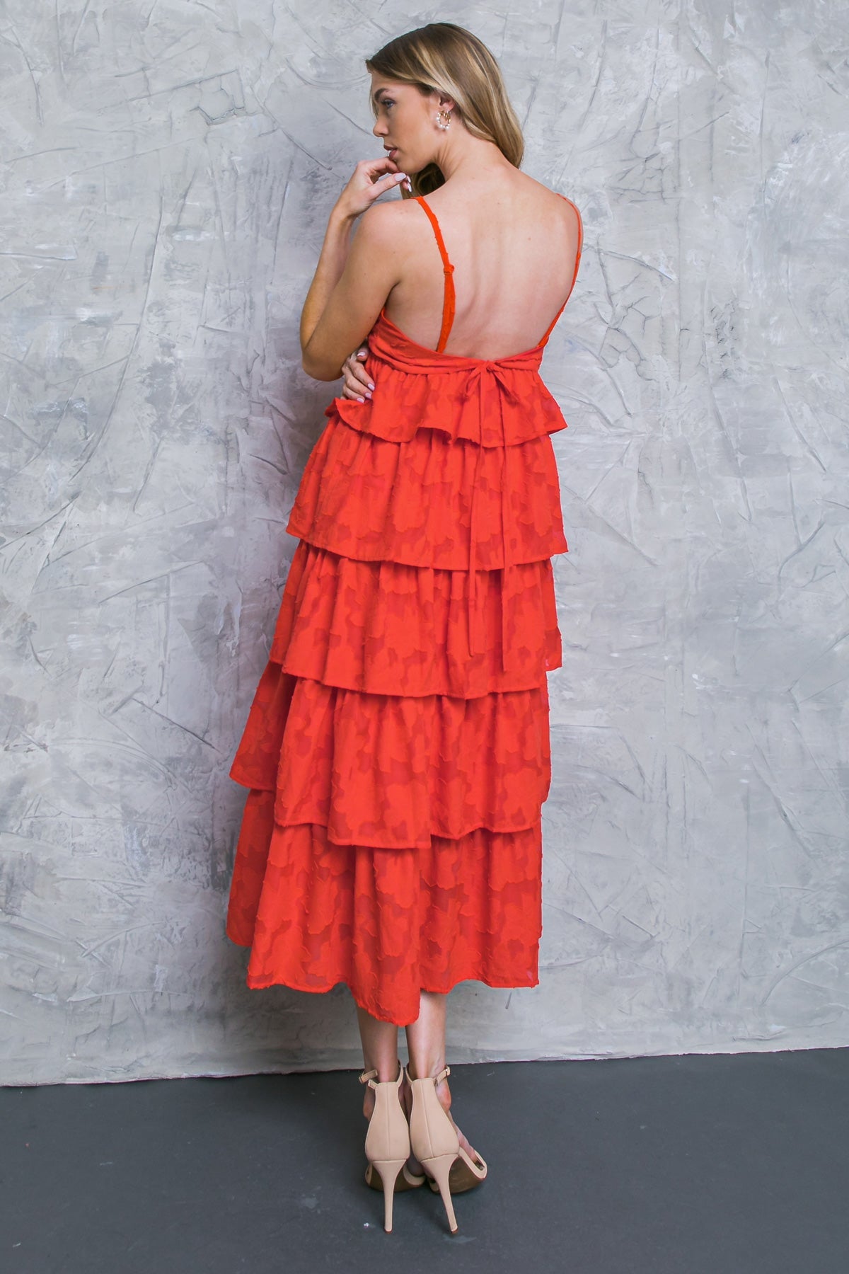 A woman, dressed in a tiered red dress, stands with her back turned. She touches her chin, facing a textured gray wall in a minimalist setting, showcasing her outfit and posture.