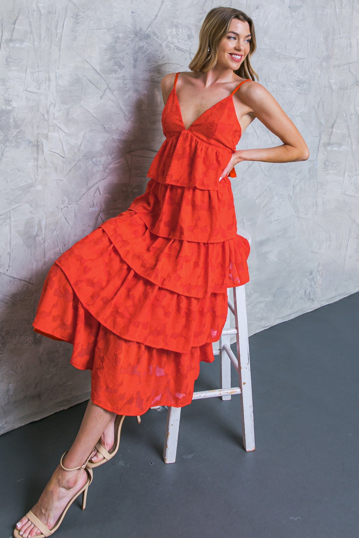 A woman in a tiered red dress poses on a white stool, smiling confidently. The background features a textured gray wall, with a dark floor complementing her stylish outfit and heels.