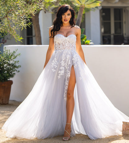 A woman poses elegantly in a flowing, light gray gown with floral embroidery and a thigh-high slit. She stands outdoors, surrounded by greenery and a sunlit, sandy area.