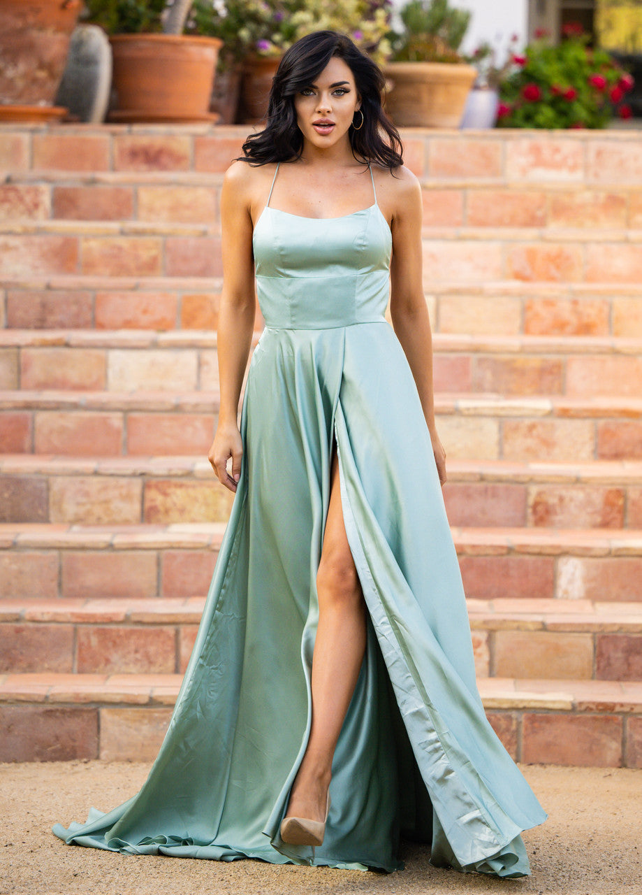 A woman in a flowing light green gown with a thigh-high slit stands on stone steps, confidently posing with a slight smile. She wears beige heels and hoop earrings.