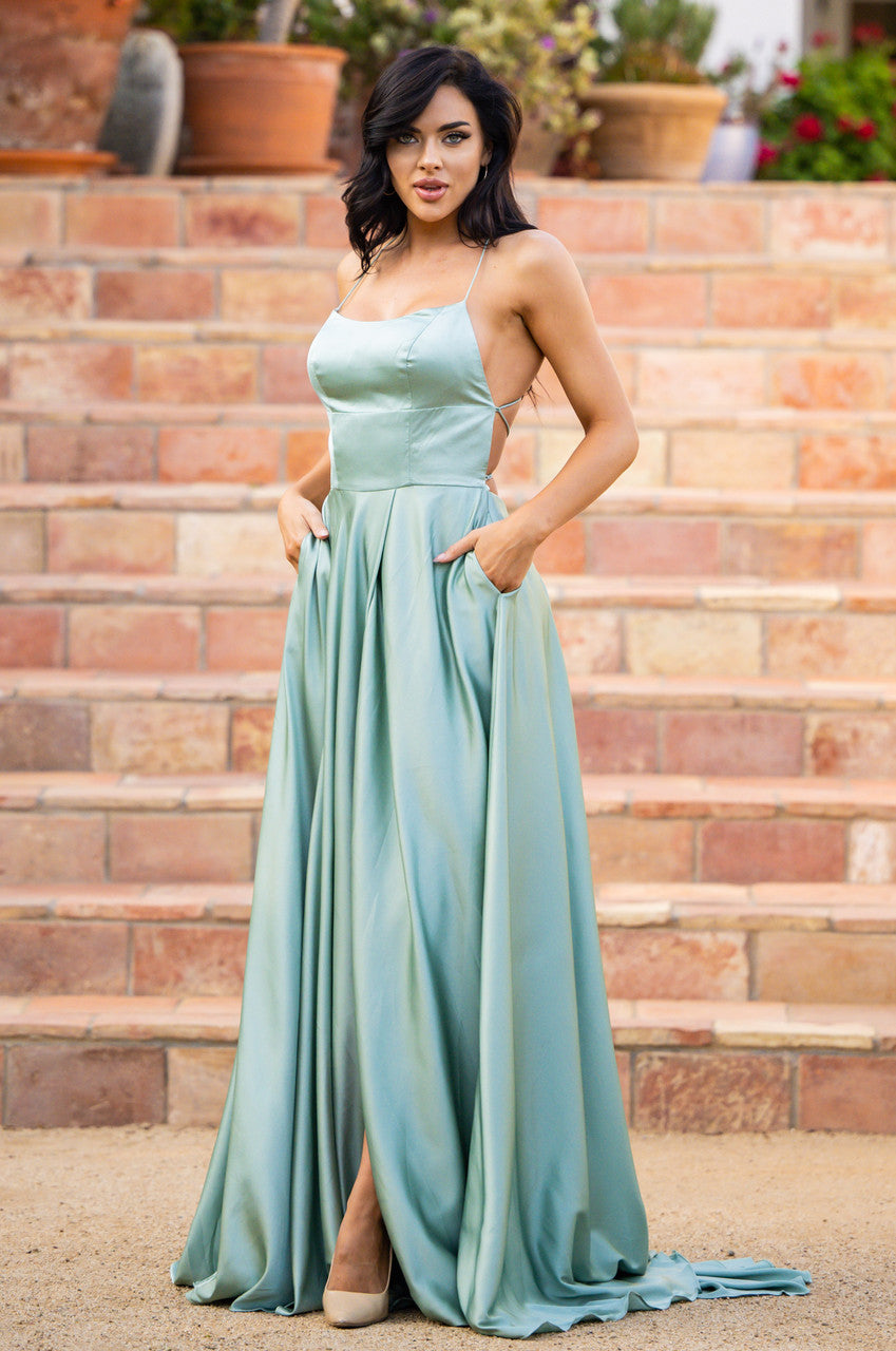 A woman in a long, silky mint green dress stands with her hands in the pockets. She poses confidently on stone steps, surrounded by potted plants and a garden setting.