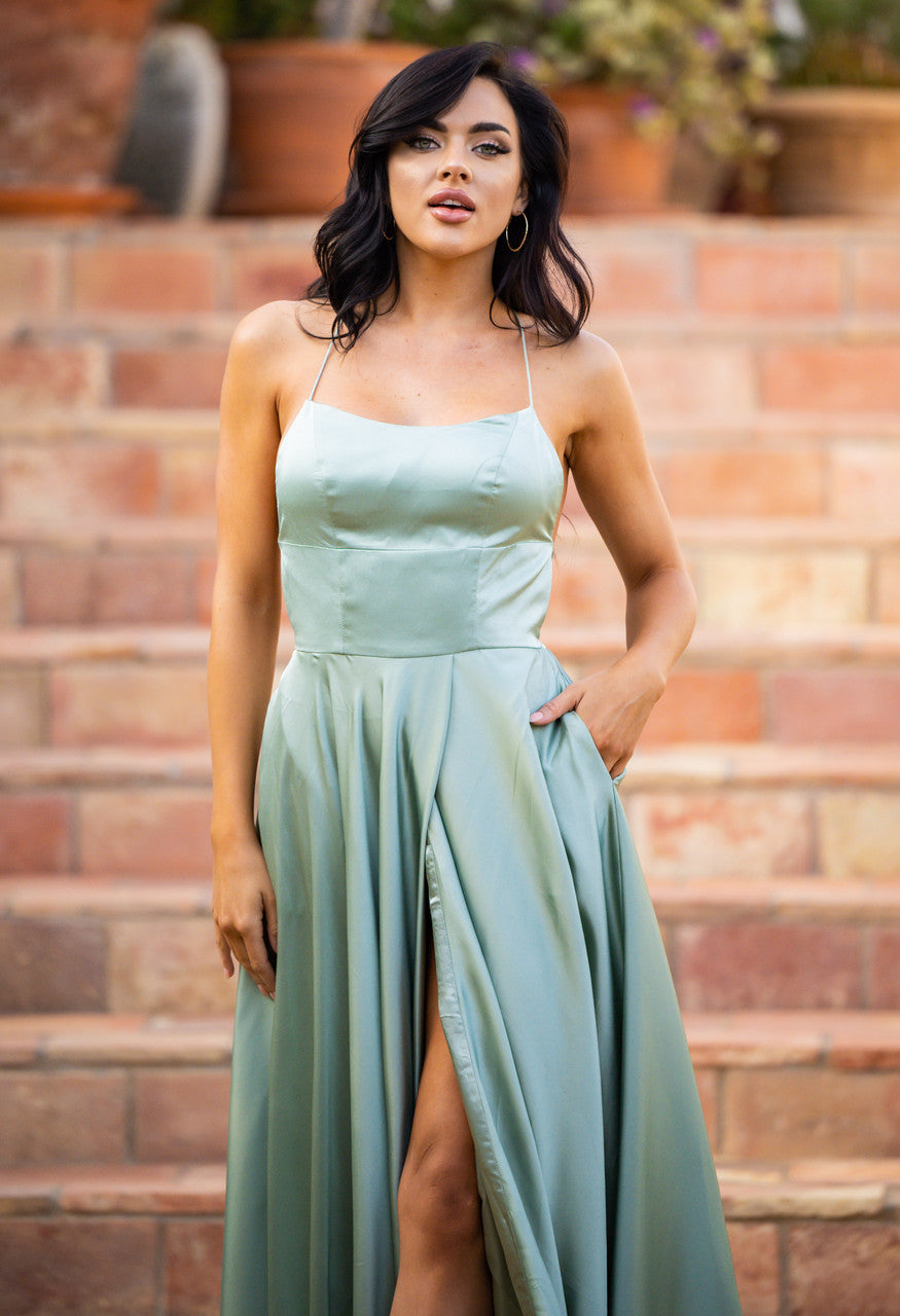 A woman in a light green, sleeveless gown stands confidently on stone steps, with her hand resting in the dress's pocket, surrounded by lush plants and warm-colored stone.