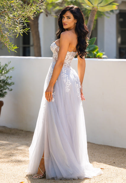 A woman in a flowing, light gray gown stands outdoors, facing slightly back. She has long, dark hair and is surrounded by greenery and a white wall in a sunny environment.