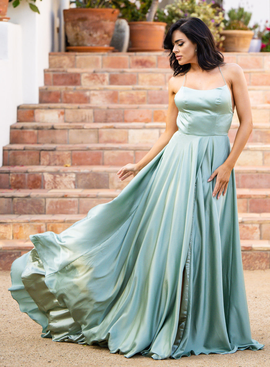 A woman in a flowing, light green gown gracefully stands on steps, gently adjusting the fabric as it billows around her, surrounded by potted plants and a serene outdoor setting.