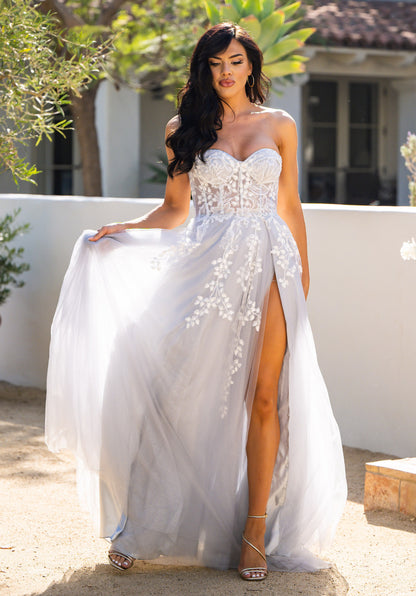 A woman in a flowing, light gray gown stands gracefully, lifting the fabric with one hand. Sunlight filters through surrounding greenery, highlighting her pose in a serene outdoor setting.