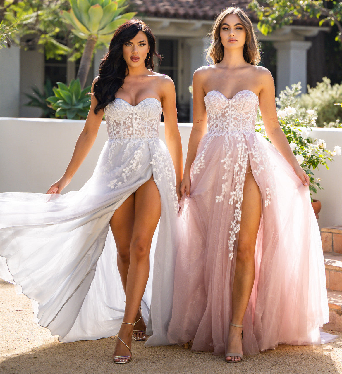 Two women walk forward, wearing elegant, flowing dresses in gray and pink, adorned with floral embroidery. They are in a sunny outdoor setting with greenery and a building in the background.