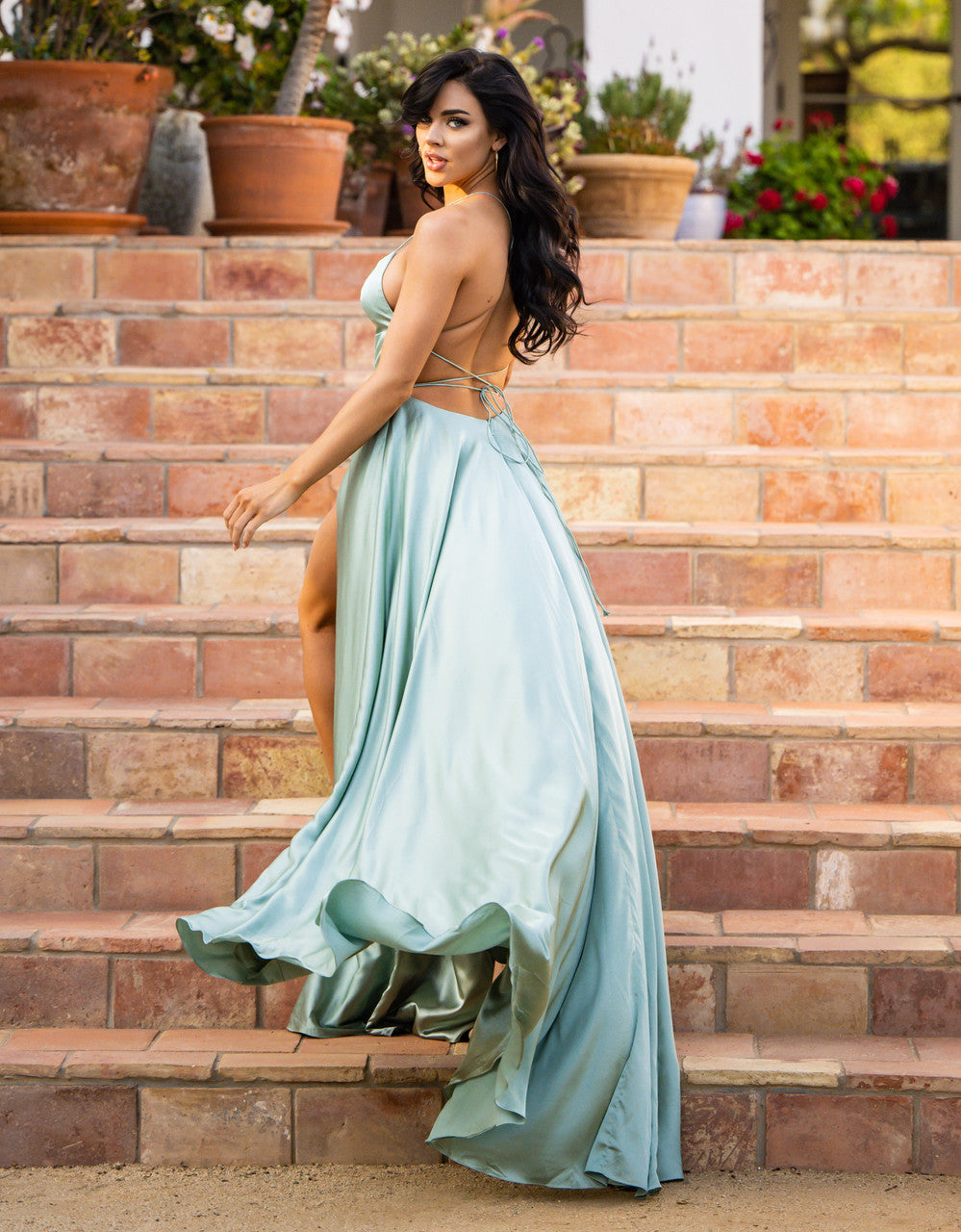 A woman in a flowing light blue gown walks down a set of stone steps, surrounded by potted plants, radiating elegance and confidence. Her dark hair flows softly behind her.