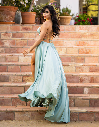 A woman in a flowing light blue gown walks down a set of stone steps, surrounded by potted plants, radiating elegance and confidence. Her dark hair flows softly behind her.