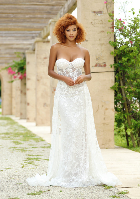 A woman stands confidently in a white lace gown, hands clasped in front. She is surrounded by stone pillars and greenery, with subtle floral accents in the background.