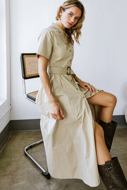 A woman in a light beige dress is seated on a metallic chair, her right arm resting on her thigh. She gazes toward the camera, surrounded by a minimalistic interior.