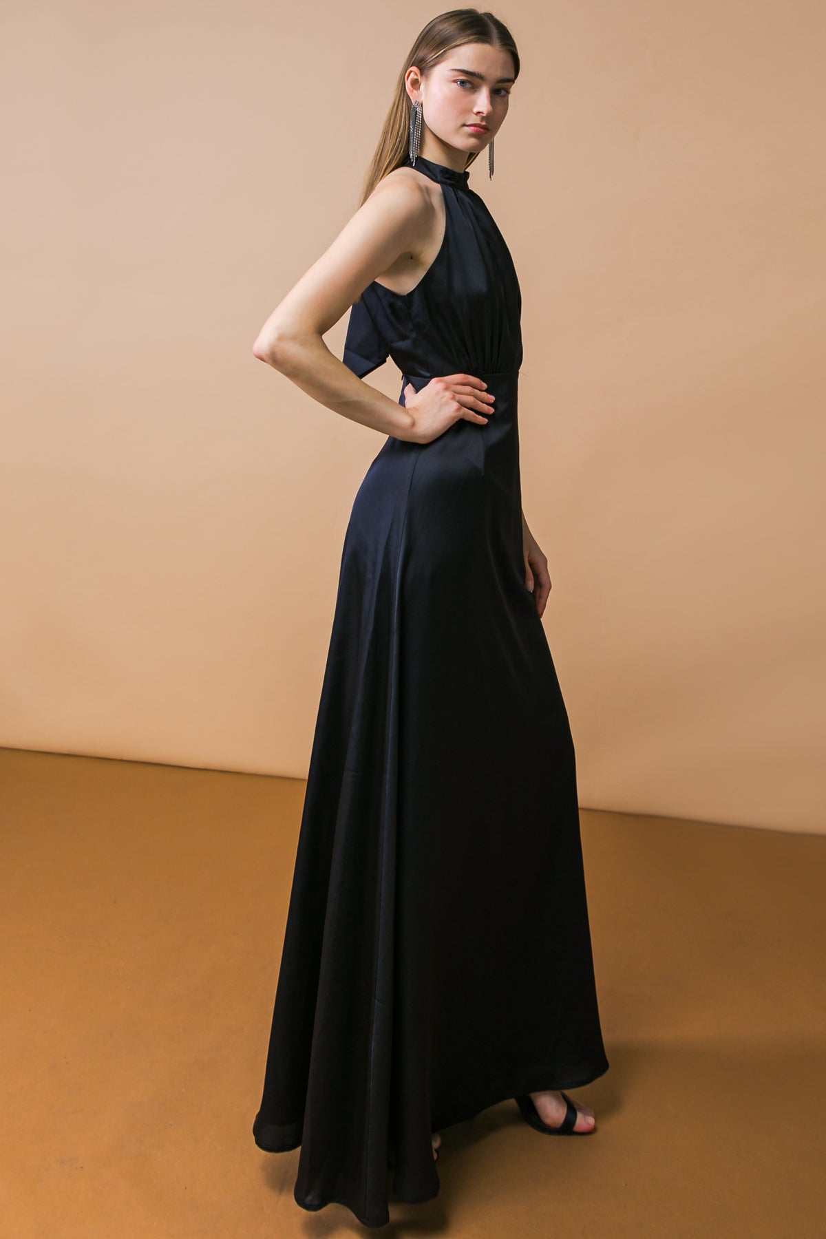 A model wears a sleek, black evening gown with a halter neck and flowing skirt, striking a pose against a soft, beige background, showcasing elegance and style.