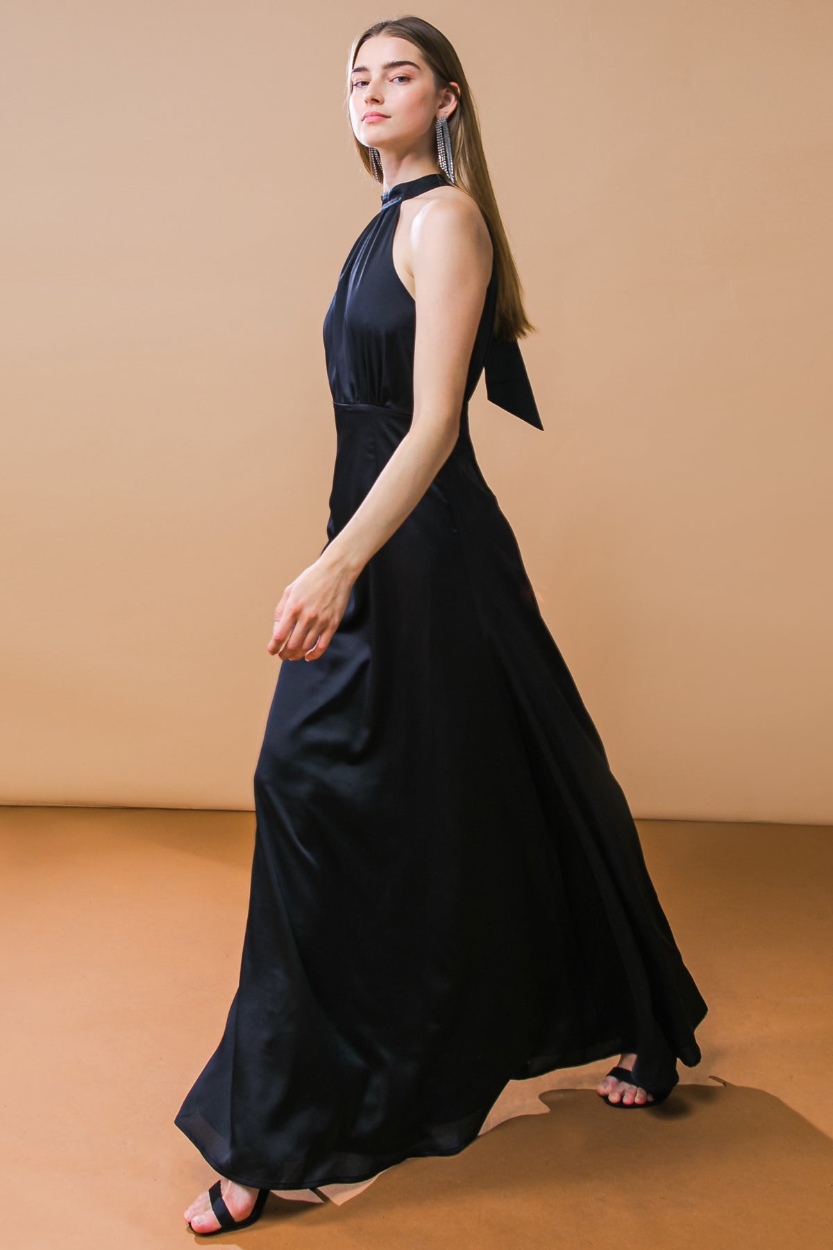 A woman in a flowing black dress walks gracefully across a smooth, warm-toned floor, showcasing elegance against a soft beige background, complemented by her long hair and statement earrings.