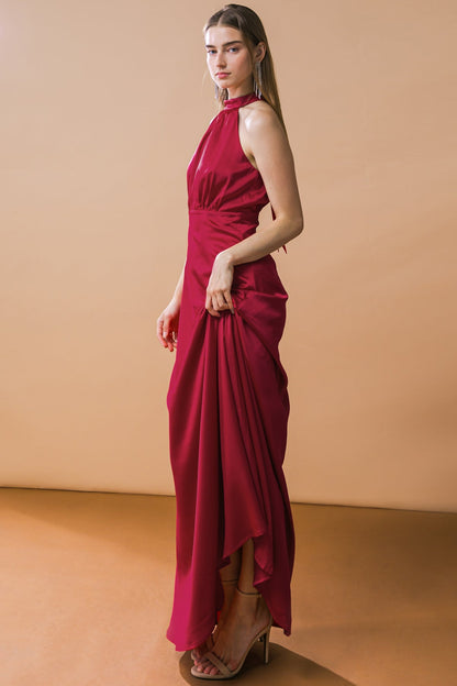 A woman stands elegantly in a deep red gown, holding the fabric gently. The backdrop is a soft beige, highlighting the dress and her poised demeanor against a simple, clean environment.