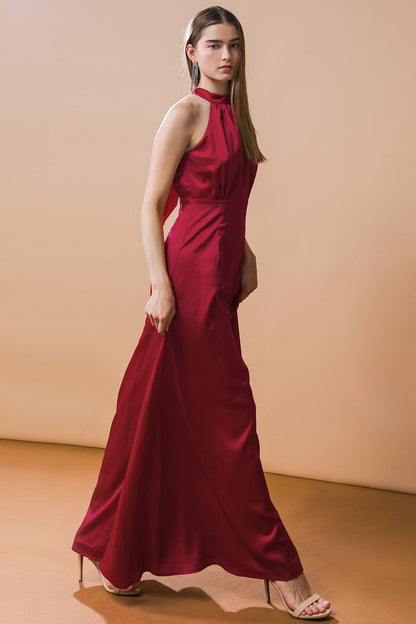 A woman in a flowing, sleeveless red gown walks gracefully, exuding elegance. She stands against a smooth beige backdrop, highlighting the dress and her poised demeanor.