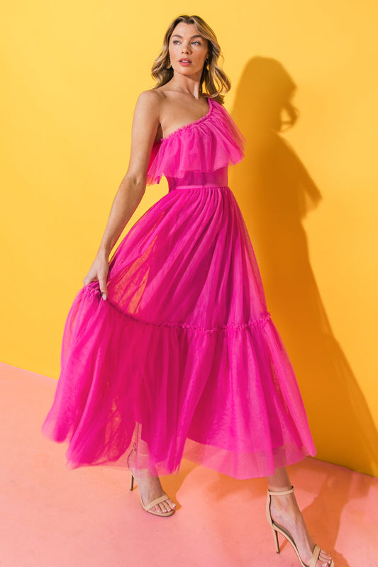 A woman wears a flowing, one-shoulder pink dress, gracefully lifting the fabric while standing against a vibrant yellow backdrop. She pairs the dress with nude high-heeled sandals.