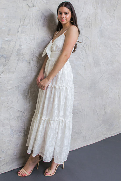 A young woman stands gracefully against a textured light gray wall, wearing a flowing white maxi dress with a bow detail at the neckline, complemented by nude high-heeled sandals.