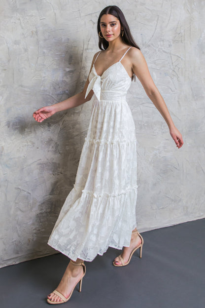 A woman wears a tiered, white dress with a bow detail, walking gracefully against a textured, light gray wall. She pairs the dress with beige high-heeled sandals.