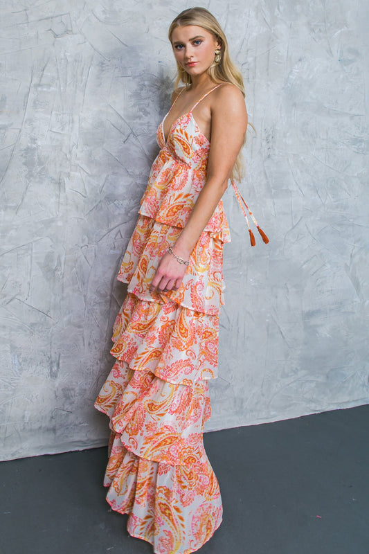 A woman stands gracefully in a layered, colorful dress adorned with paisley patterns. She poses against a textured gray backdrop, showcasing her elegant style and confidence.