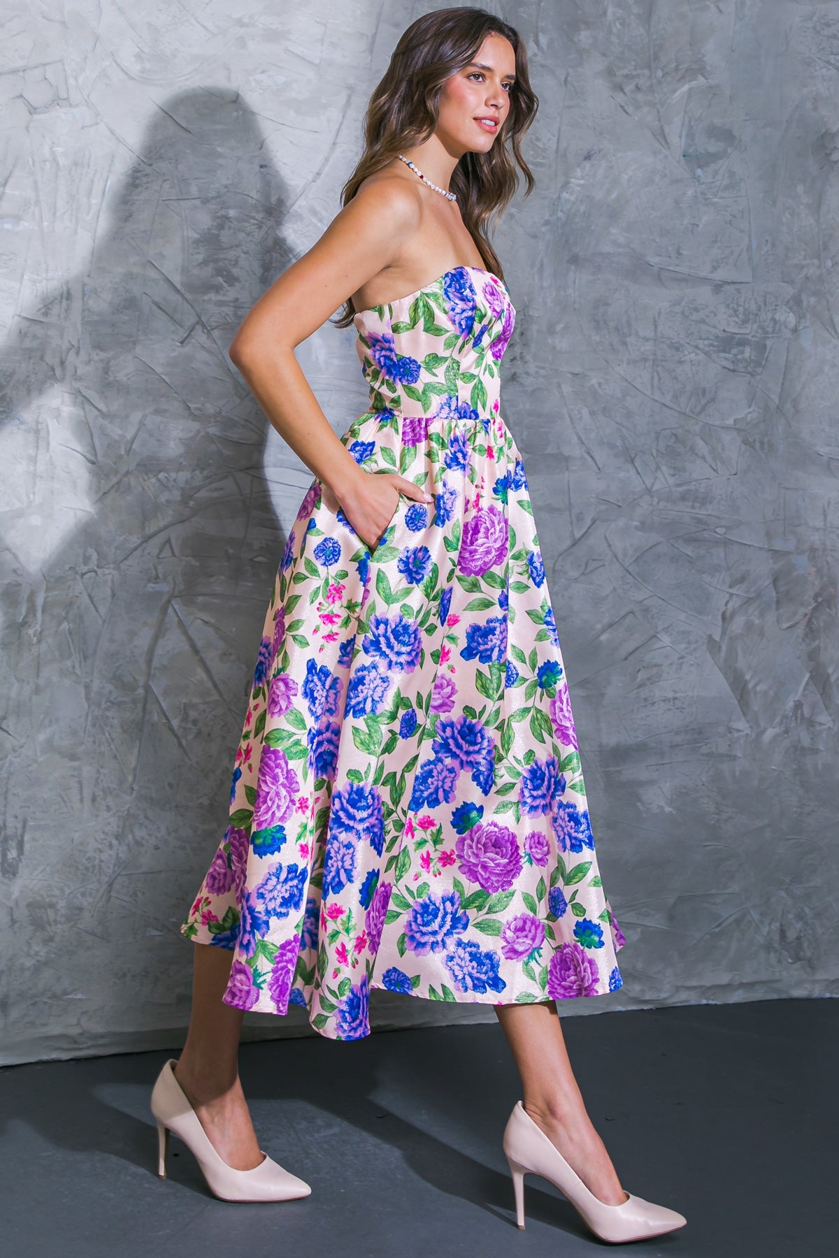 A woman wearing a floral strapless dress walks confidently, with one hand in her pocket. The backdrop features a textured gray wall, enhancing her elegant pose and outfit.