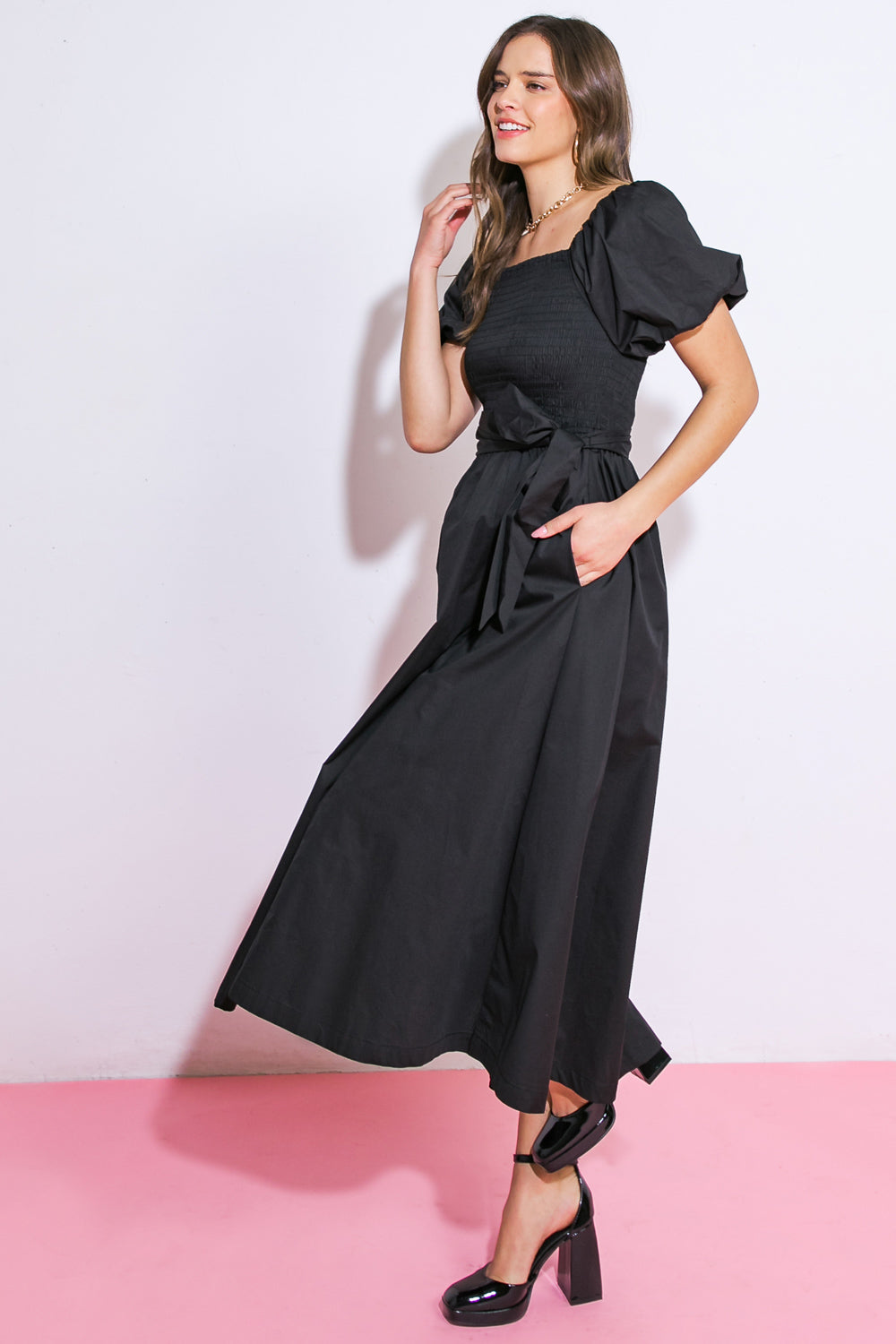 A woman with wavy hair wears a black dress with puff sleeves and a belt. She poses confidently, touching her hair, against a white wall and pink floor.