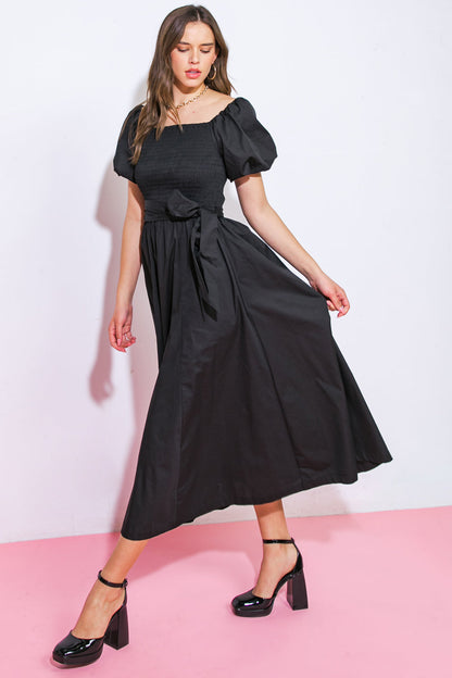 A woman wearing a black, puff-sleeve dress with a fitted bodice and flowing skirt stands gracefully, moving one hand to the side. The backdrop features a white wall and pink floor.