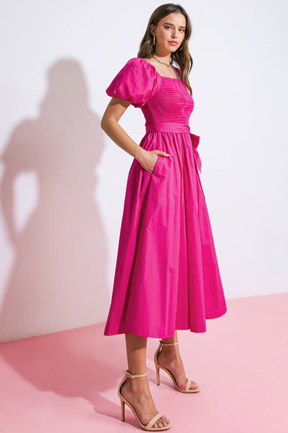 A woman stands elegantly in a bright pink dress with puff sleeves, pocketed skirt, and a tie at the waist, set against a light-colored wall and pink floor.