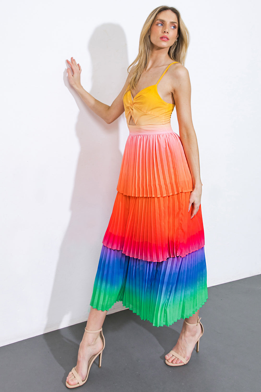 A woman stands beside a white wall, wearing a vibrant, multi-colored pleated skirt that fades from orange to green, paired with a yellow top, showcasing a stylish pose.