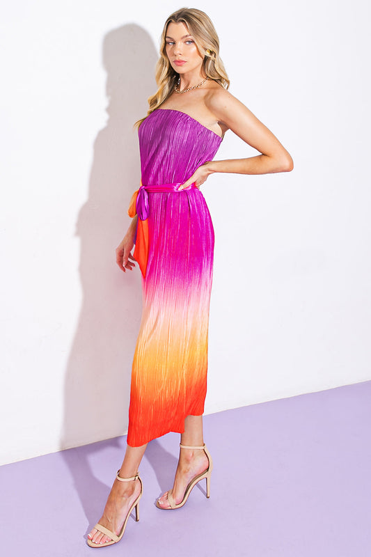 A woman stands confidently in a colorful, pleated, strapless dress that transitions from purple to orange. She poses against a white wall and purple floor, wearing stylish sandals.