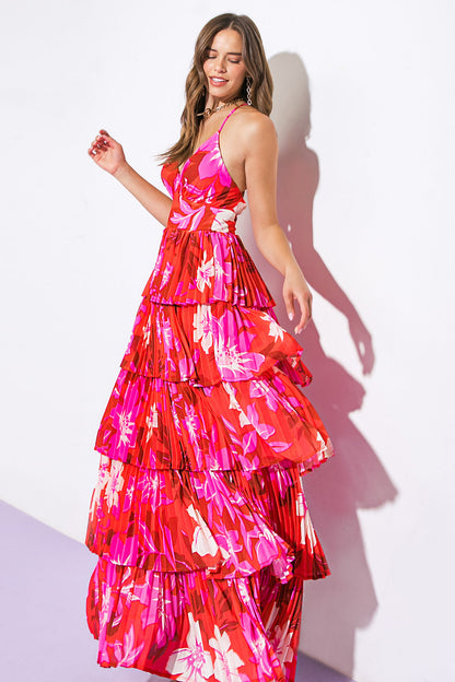 A woman is wearing a vibrant, tiered red dress with pink floral patterns. She stands gracefully against a white wall, smiling and slightly raising one hand, exuding a joyful atmosphere.