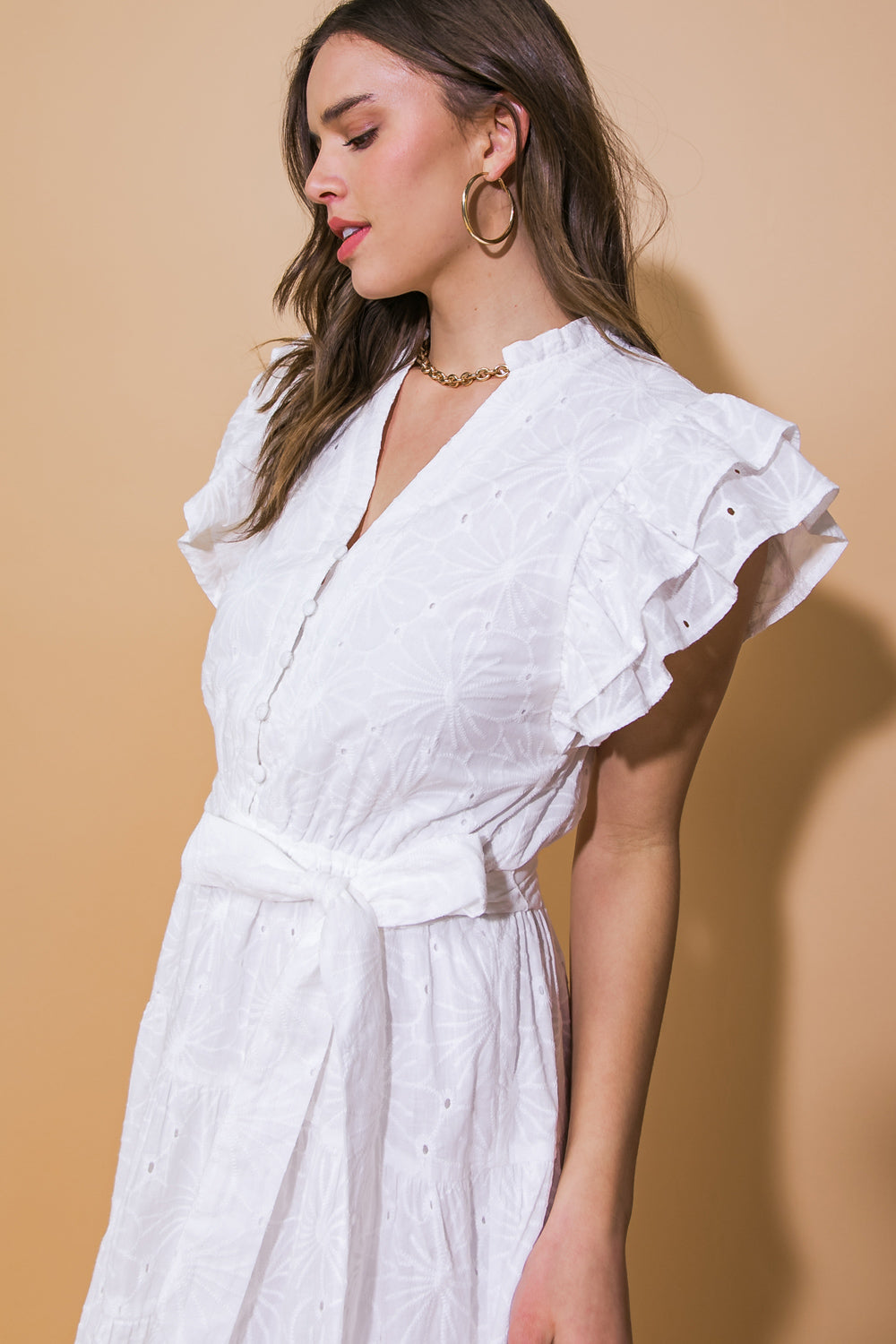 A woman wears a white, ruffled dress with a belt, facing to the side. She has gold hoop earrings and a chain necklace, against a warm, beige background.