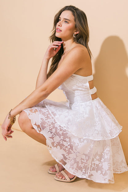 A woman in a white floral dress crouches gracefully on one knee, thoughtfully touching her face, against a soft beige background that enhances her elegant pose.