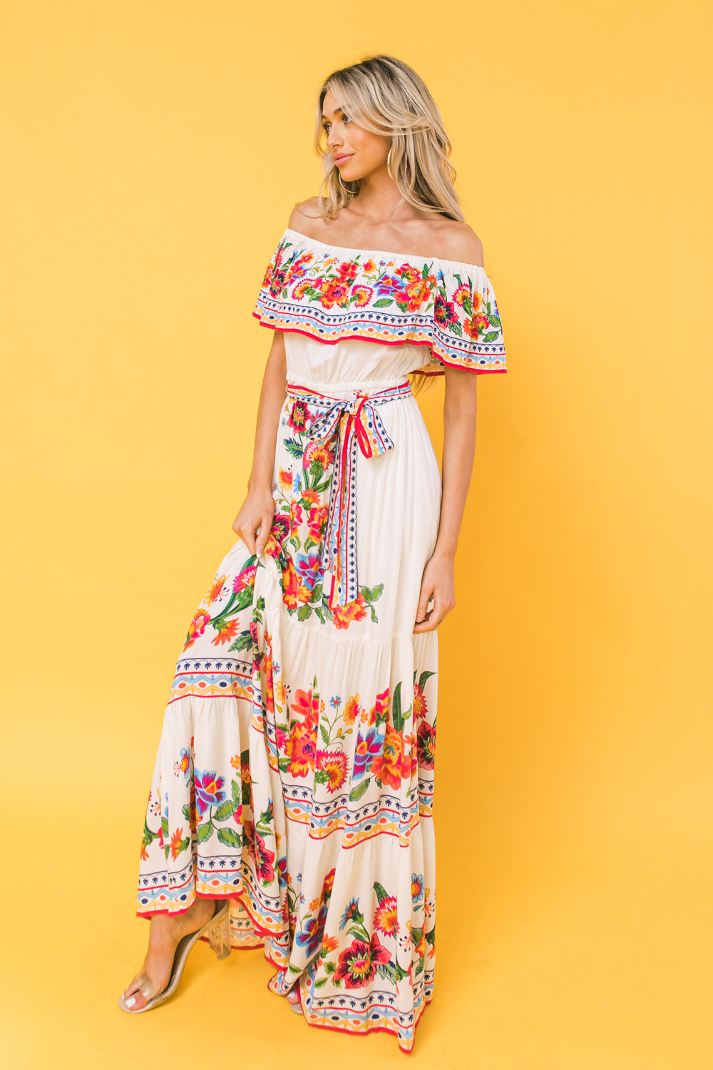 A woman stands gracefully wearing a long, off-the-shoulder white dress adorned with vibrant floral patterns and a colorful belt, against a bright yellow backdrop.