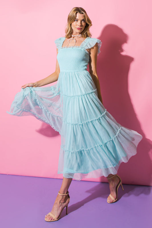 A woman in a light blue, flowy dress with ruffles twirls playfully, creating movement. She's against a pink backdrop, with a purple floor, showcasing a vibrant, stylish atmosphere.