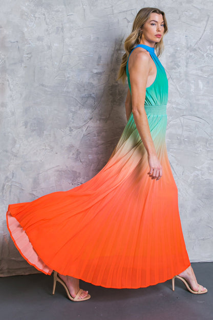 A model walks gracefully in a flowing, ombre maxi dress, transitioning from green at the top to orange at the hem, against a textured gray wall.