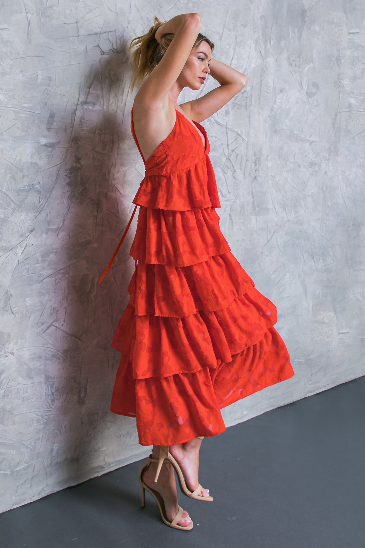 A woman stands against a textured gray wall, adjusting her hair. She wears a bright red, tiered dress and nude high-heeled sandals, creating a striking contrast in the setting.