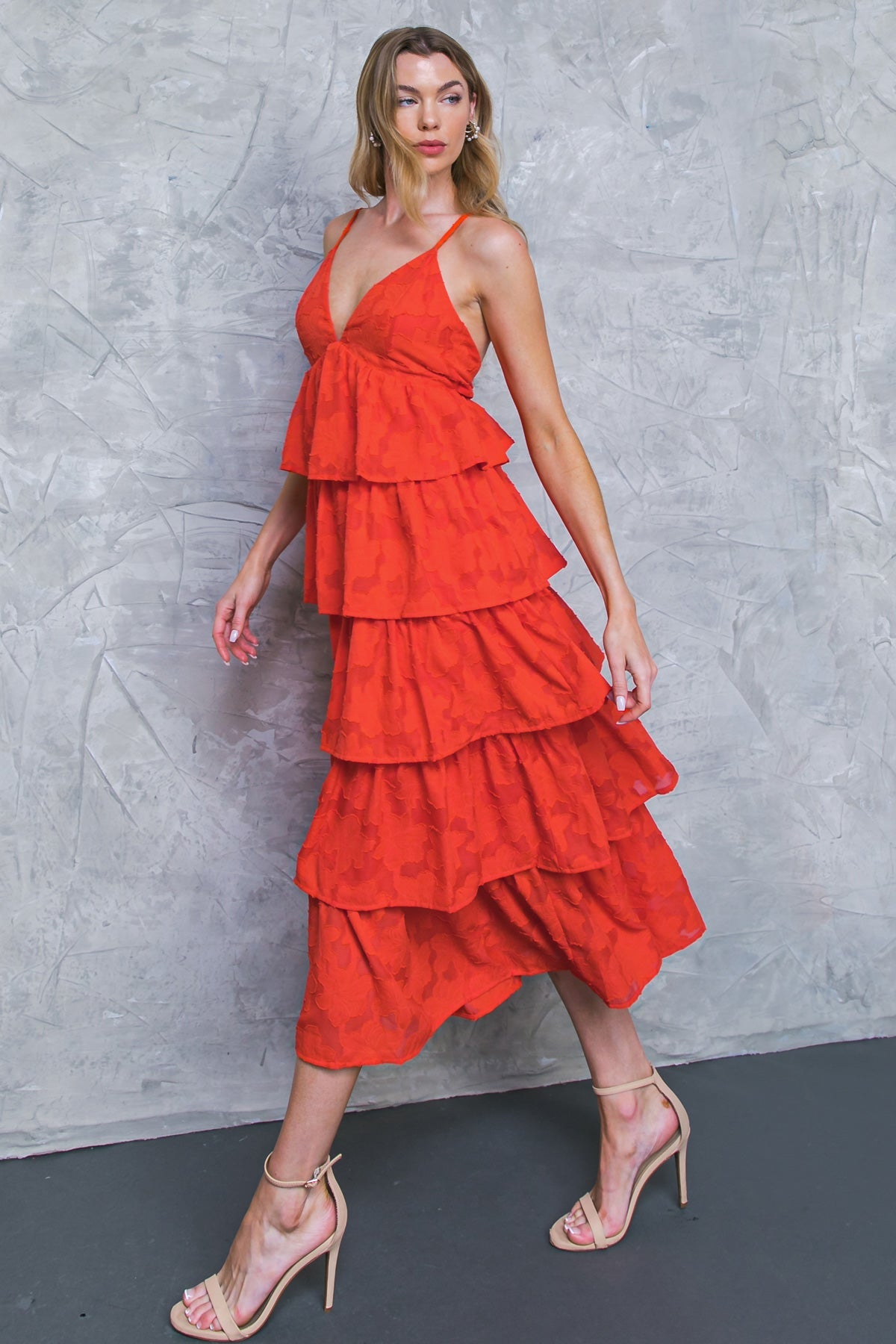 A model walks gracefully, wearing a vibrant red, tiered dress that flows around her. She stands against a textured gray wall, showcasing her high-heeled sandals.