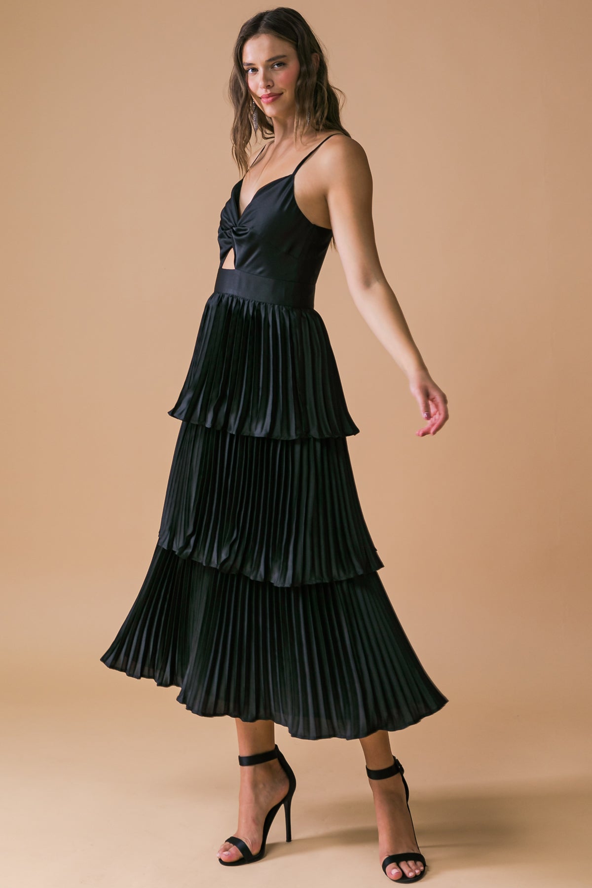 A model wears a black, pleated midi dress with a twist detail at the chest. She poses gracefully against a warm, beige background, showcasing the dress's flow and elegance.