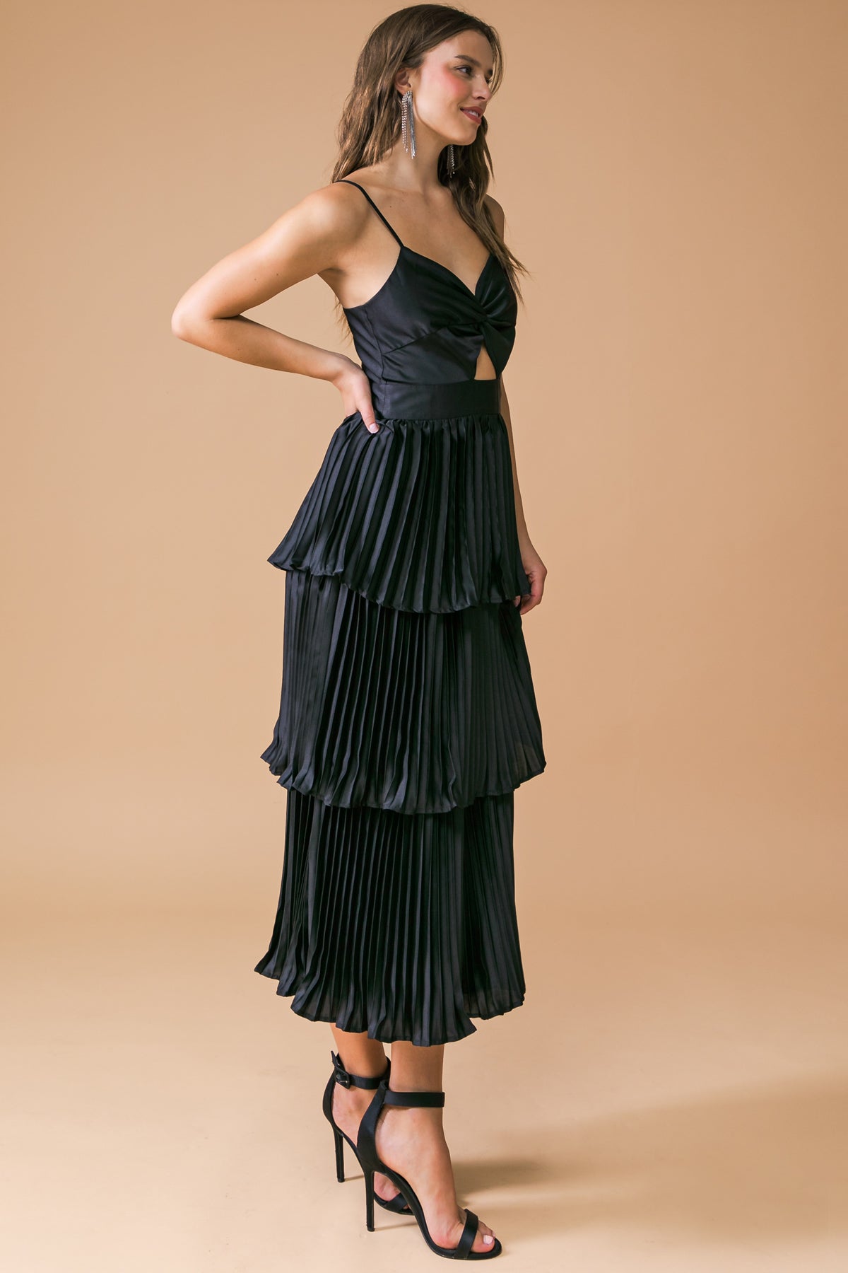 A model wearing a black, pleated dress with a fitted upper and layered skirt stands in profile. She accessorizes with long earrings against a plain beige background.