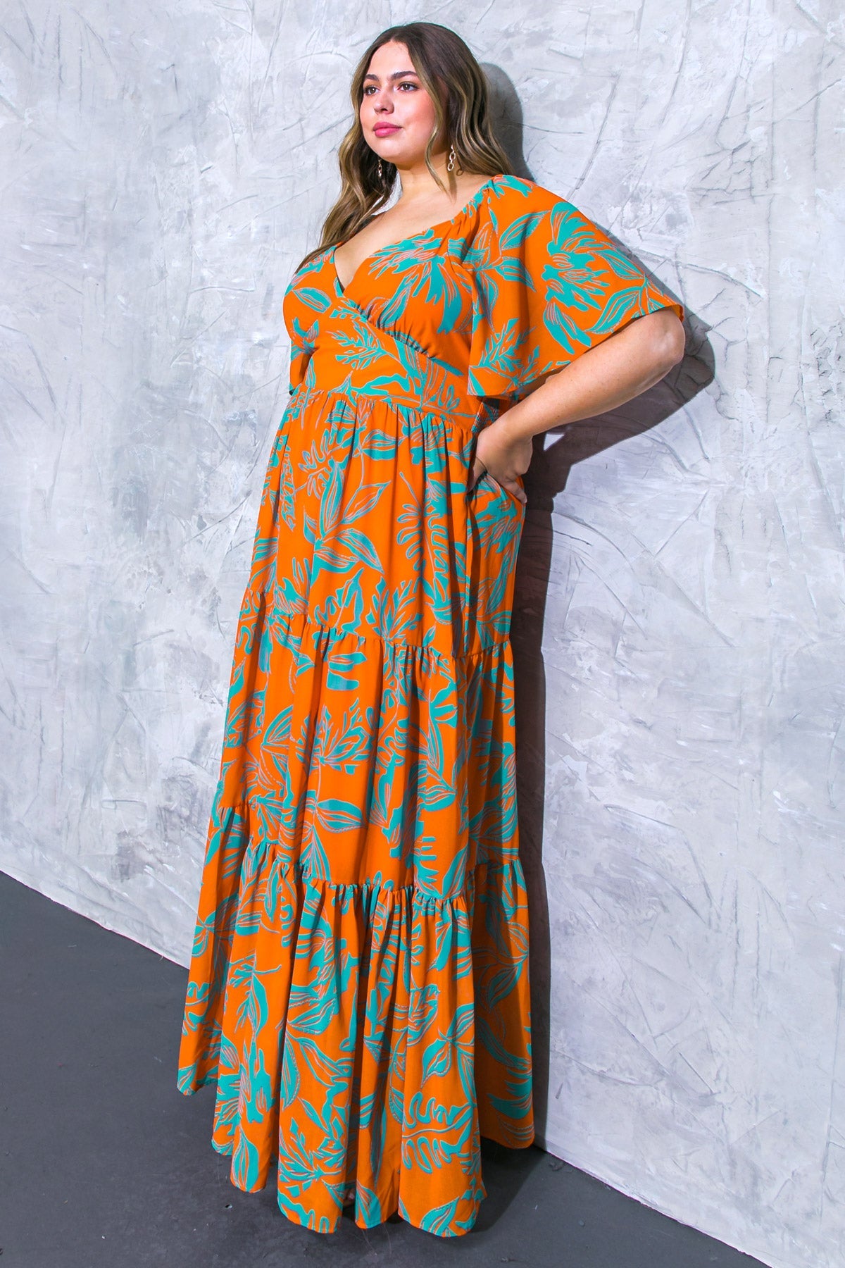 A woman stands confidently against a textured gray wall, wearing a vibrant orange maxi dress adorned with teal botanical patterns, displaying a relaxed, elegant pose with her hands in pockets.